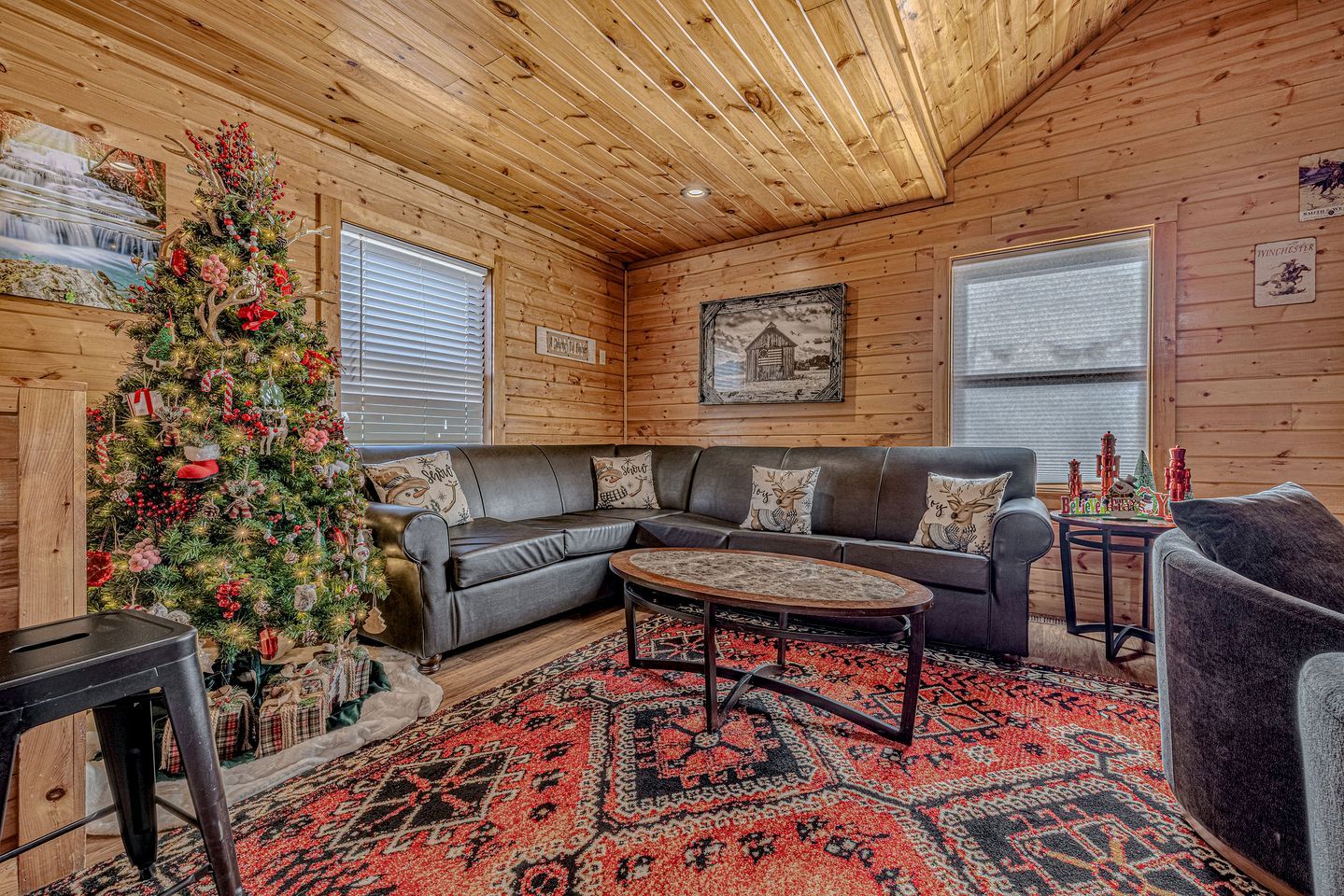 A cozy cabin interior with a wooden ceiling, a Christmas tree, a leather sofa, and a patterned rug.
