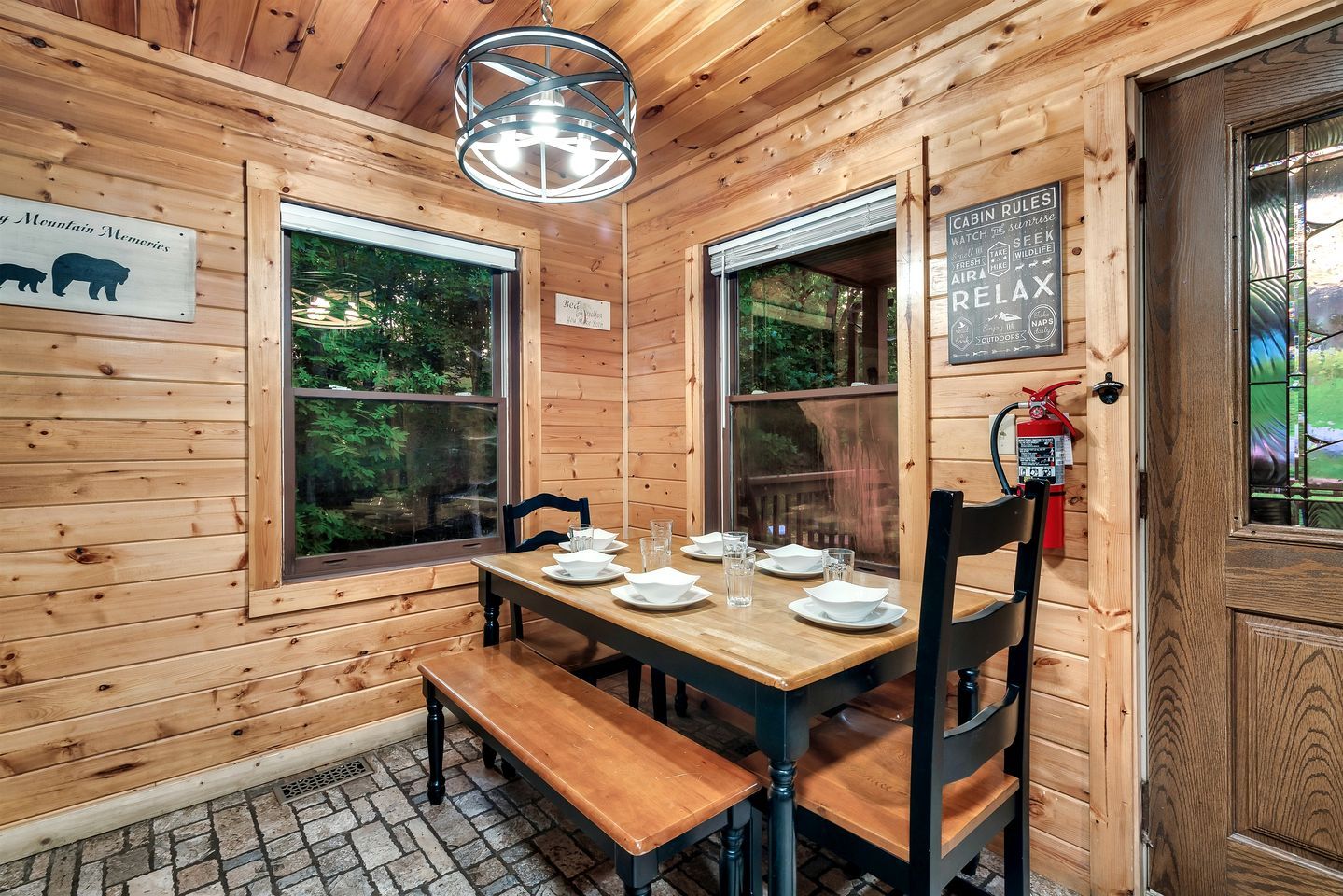Interior de una cabaña de madera con una mesa de comedor lista para cuatro comensales, dos ventanas, un extintor y un cartel con las normas de la cabaña.