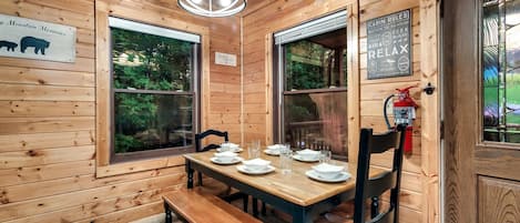 Interior de una cabaña de madera con una mesa de comedor lista para cuatro comensales, dos ventanas, un extintor y un cartel con las normas de la cabaña.