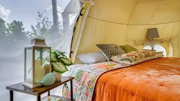 A tented room with a bed, a table with a lantern, and a window with a view of trees.