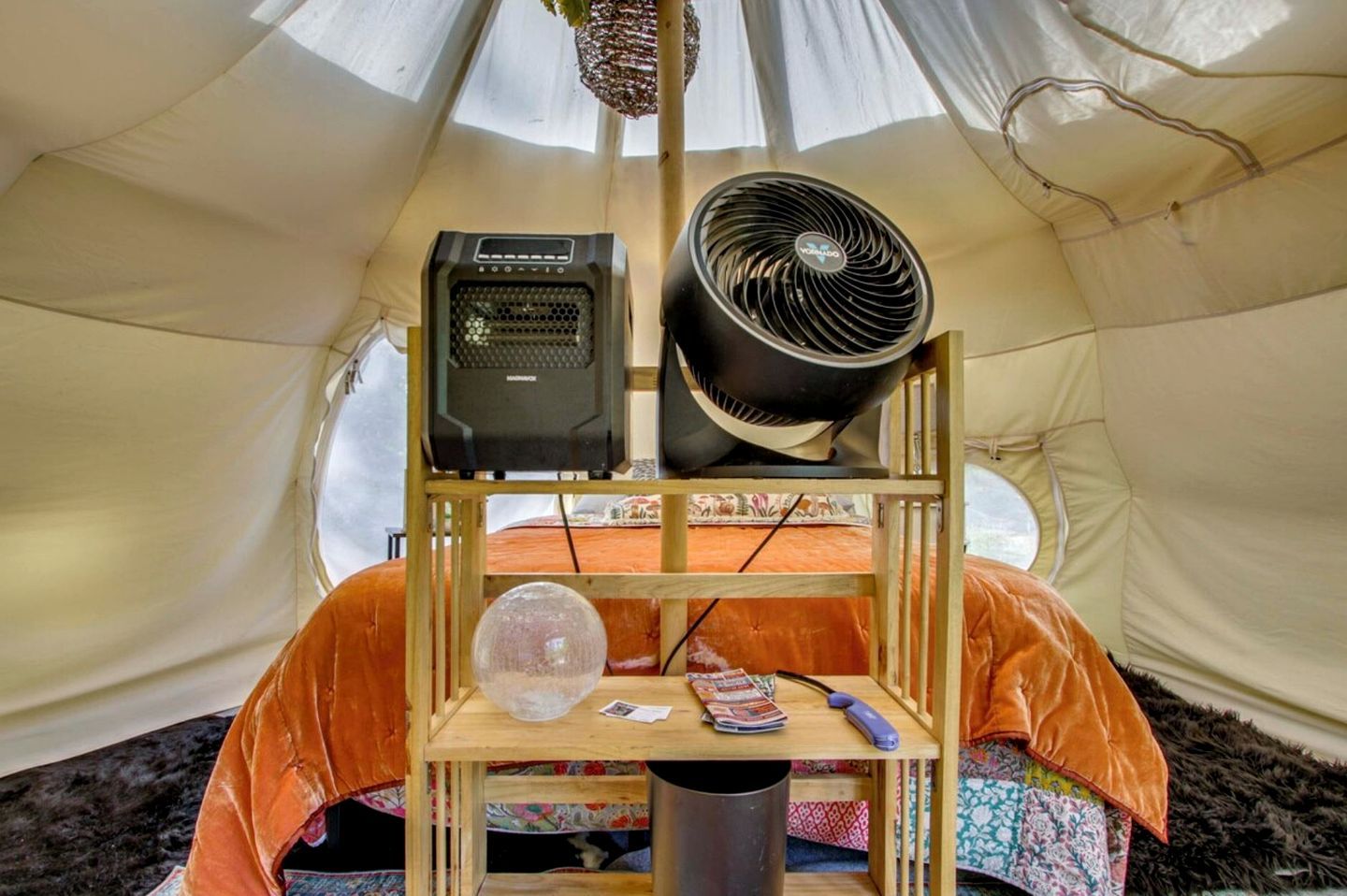 A tent interior with a bed, a speaker, a fan, a globe, and a table with a book and a remote control.