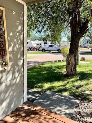 A wooden porch with a welcome sign, a tree, and a view of RVs in the background.