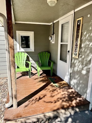 Une véranda avec deux fauteuils Adirondack verts, une terrasse en bois et une porte ornée d’une pancarte de bienvenue.