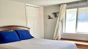 A bedroom with a wooden bed, a white bedspread, a white door, and a window with curtains.