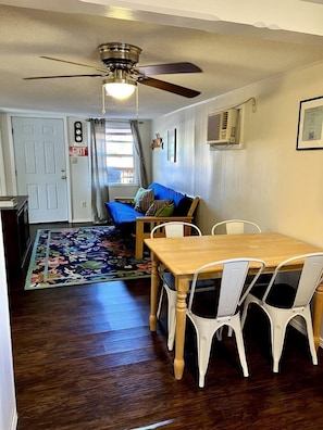 A dining area with a wooden table and white chairs, a ceiling fan, a blue sofa, and a floral rug.