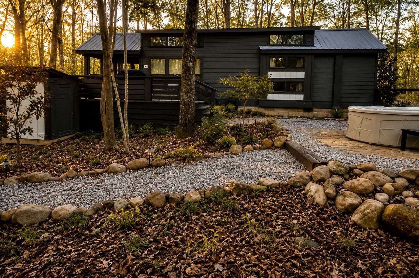 A modern cabin with a gravel driveway, surrounded by trees and a rock garden.