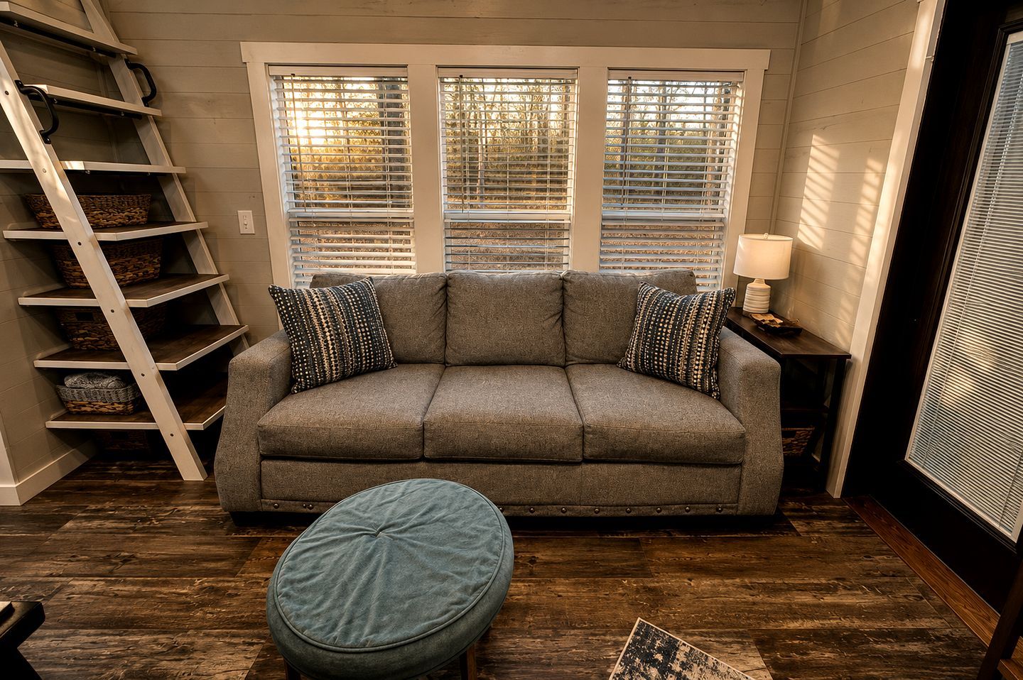 Una habitación con un sofá gris, dos almohadones estampados, un otoman redondo, una escalera de madera y una ventana con persianas.