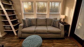 Una habitación con un sofá gris, dos almohadones estampados, un otoman redondo, una escalera de madera y una ventana con persianas.