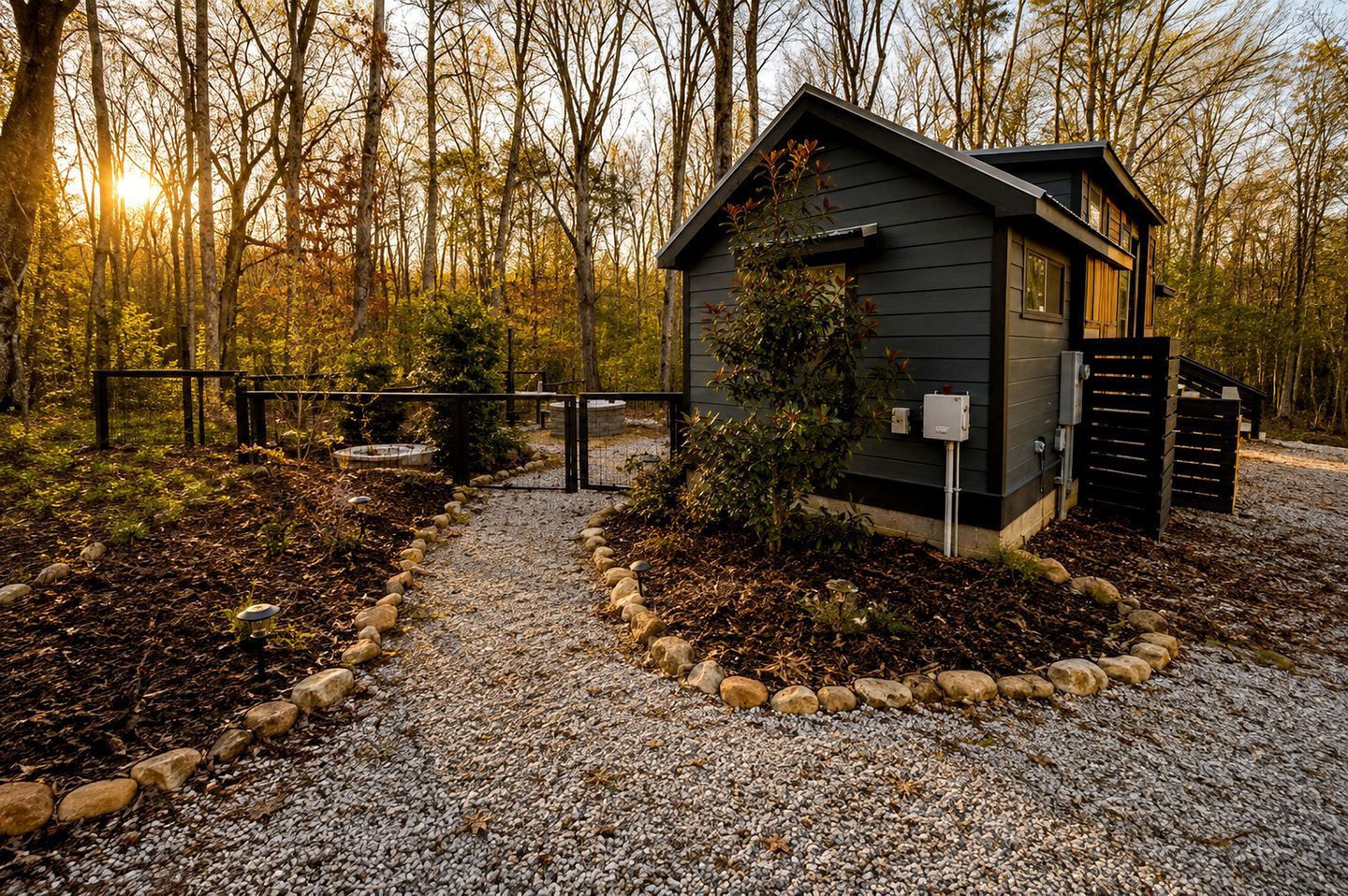 A small cabin with a gravel path leading to it, surrounded by a fence and trees.
