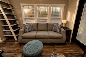 A room with a grey sofa, two patterned pillows, a round ottoman, a wooden ladder, and a window with blinds. - Chic Tiny Cabin with Jacuzzi, Screened Porch and Grill in Tennessee (Tracy City)