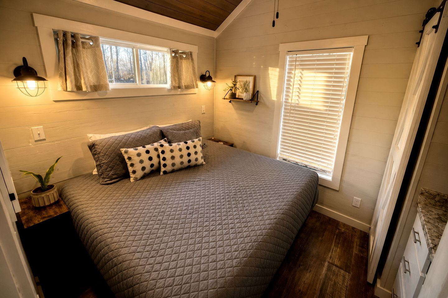 Un dormitorio con una cama grande, dos lámparas de noche, una ventana con persianas y una estantería con objetos decorativos.