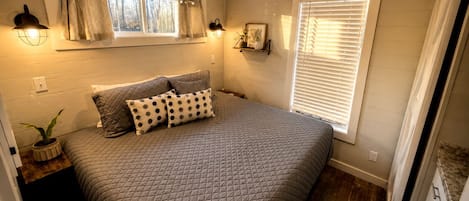 Une chambre à coucher avec un grand lit, deux lampes de chevet, une fenêtre avec des stores et une étagère ornée d’objets décoratifs.