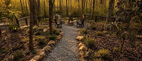 Un área boscosa con un sendero de grava que conduce a un espacio de descanso con sillas y una chimenea.