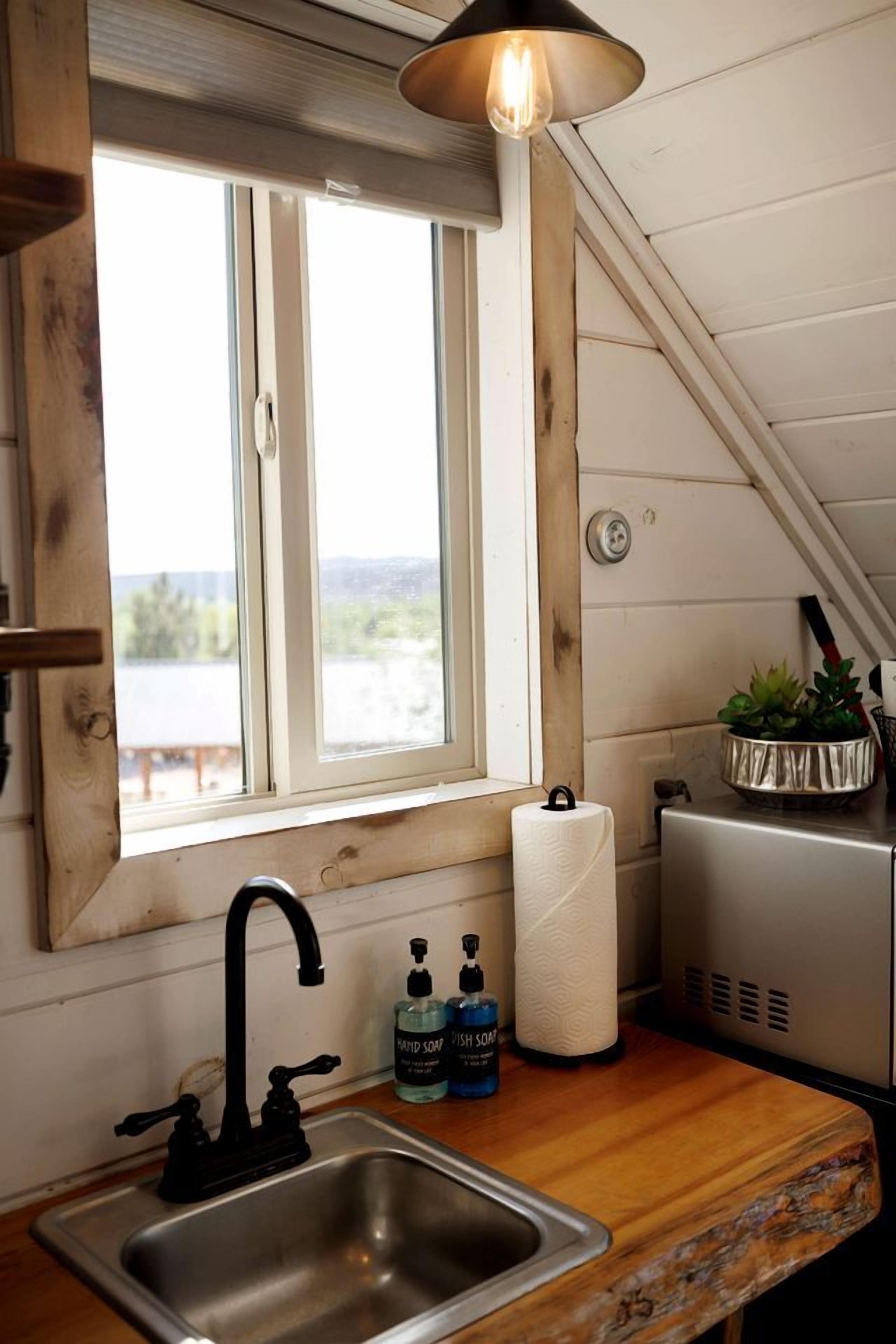 Una cocina con ventana, un lavamanos, un grifo, un dispensador de toallas y un refrigerador pequeño.