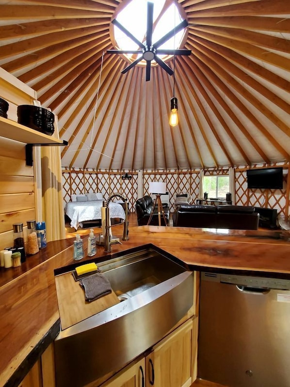 A yurt interior with a wooden ceiling, a central light fixture, a kitchen area with a sink and cabinets, and a bed with a window.