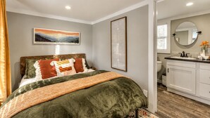 A bedroom with a bed, a bedside table, a nightstand, a window with curtains, a bathroom with a sink, mirror, and toilet. - Cozy Wooden Cabin Surrounded by Trees in Monteagle, Tennessee (Monteagle)
