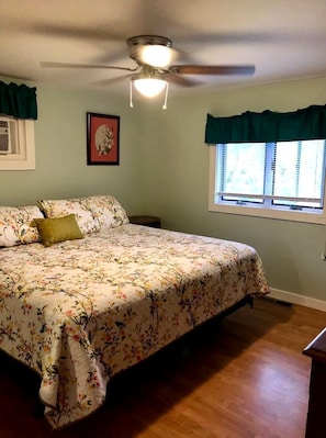 Une chambre à coucher avec un lit, un ventilateur de plafond, une fenêtre avec des rideaux et un tableau encadré au mur.