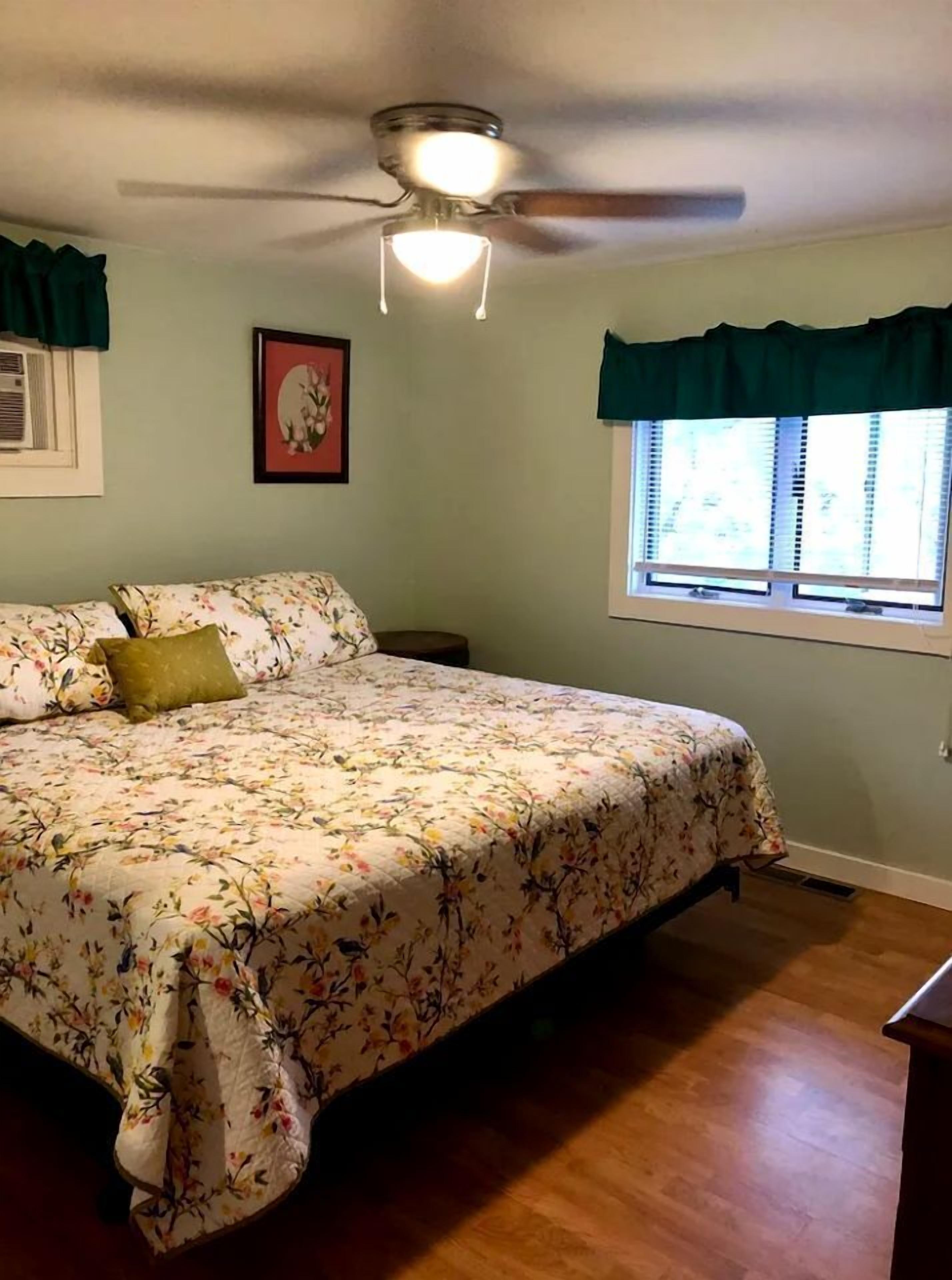 Un dormitorio con cama, ventilador de techo, ventana con cortinas e imagen enmarcada en la pared.