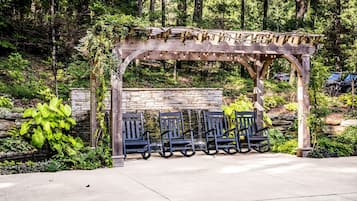 A wooden pergola with seating, surrounded by lush greenery and a stone wall.
