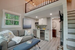 Una sala de estar moderna con un sofá gris, una mesa de comedor y una zona de cocina con armarios blancos y un microondas.