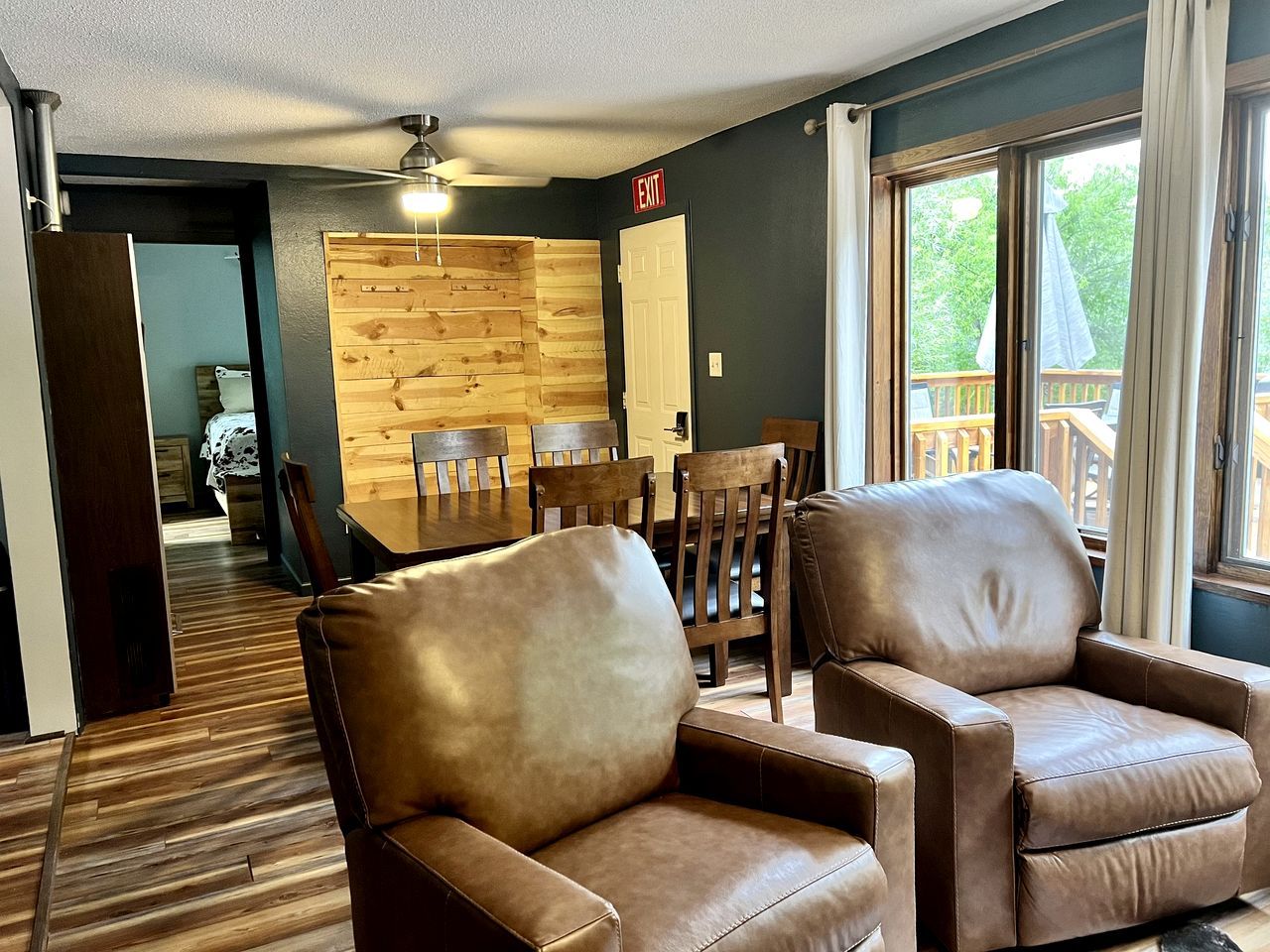 A room with a wooden wall, a dining table with chairs, a sofa, and a view through a window.