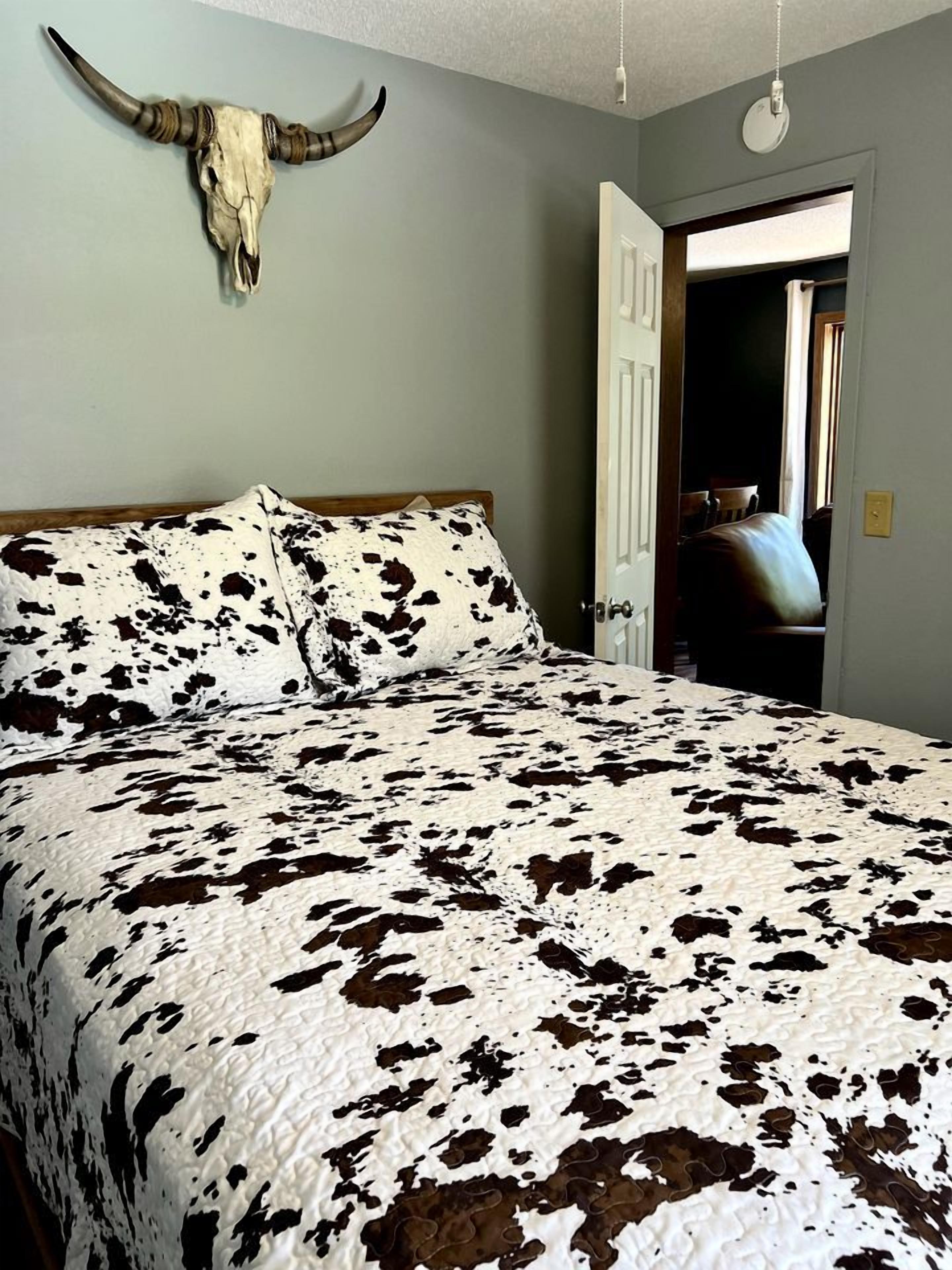 A bedroom with a cowhide patterned bedspread, a cow skull mounted on the wall, and a door leading to another room.