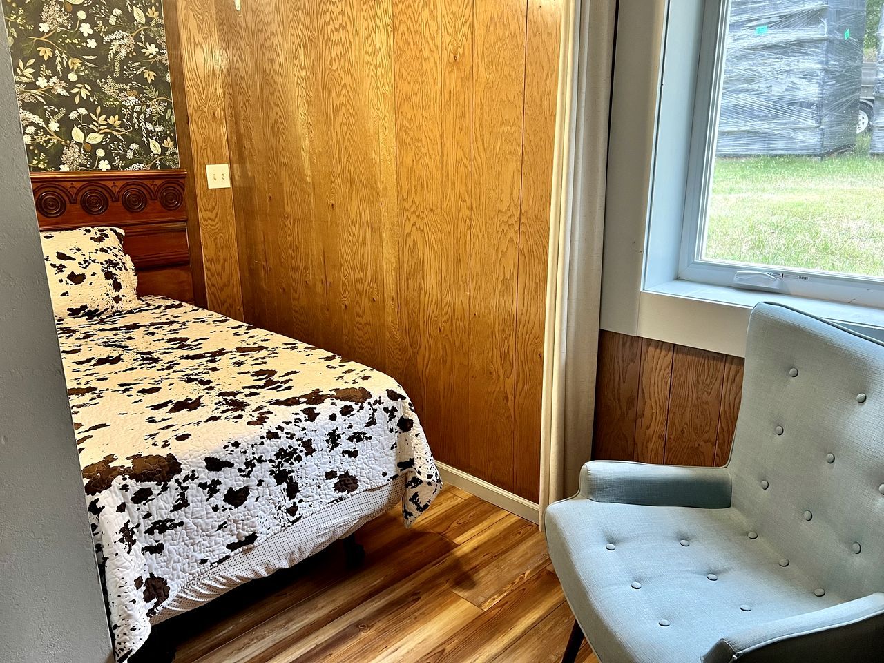 Un dormitorio con una cama, una silla, un ventanal y una puerta de madera.