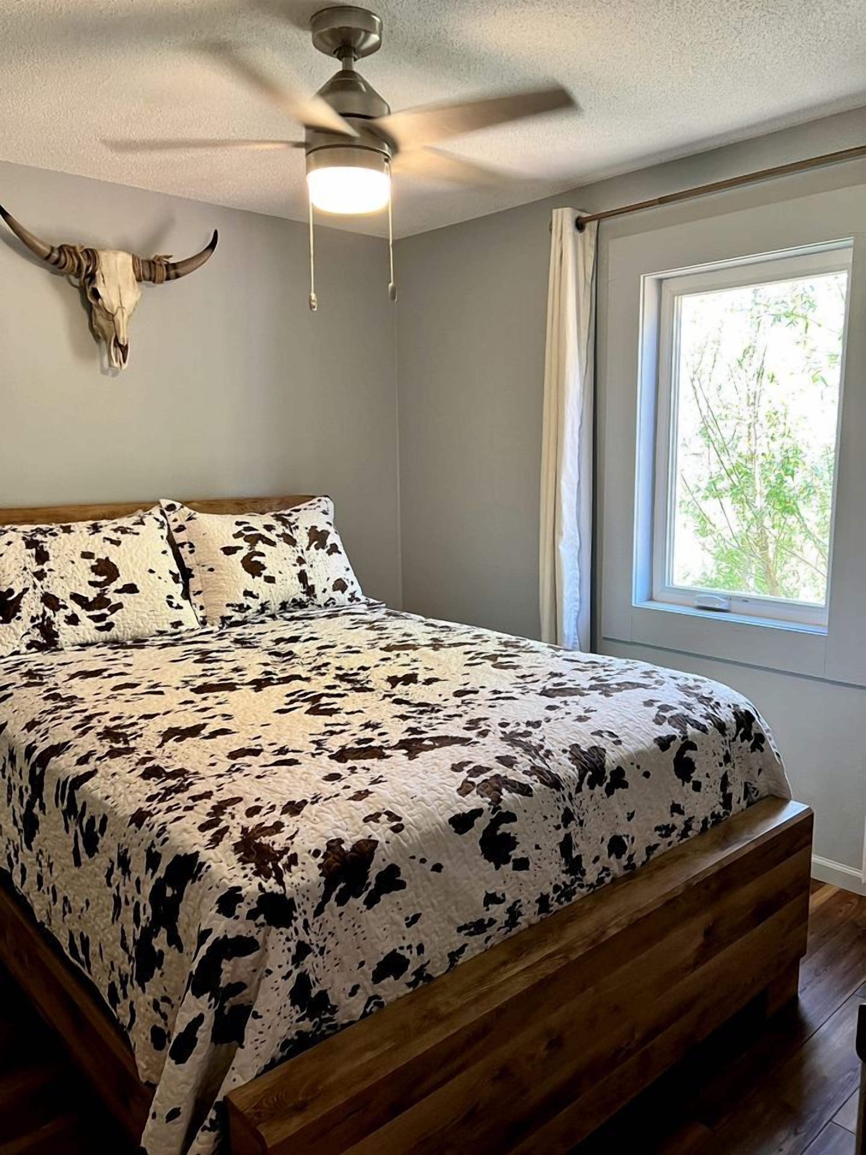 A bedroom with a cowhide-patterned bedspread, a wooden headboard, a ceiling fan, a large window with a view of greenery, and a wall-mounted cow skull decoration.