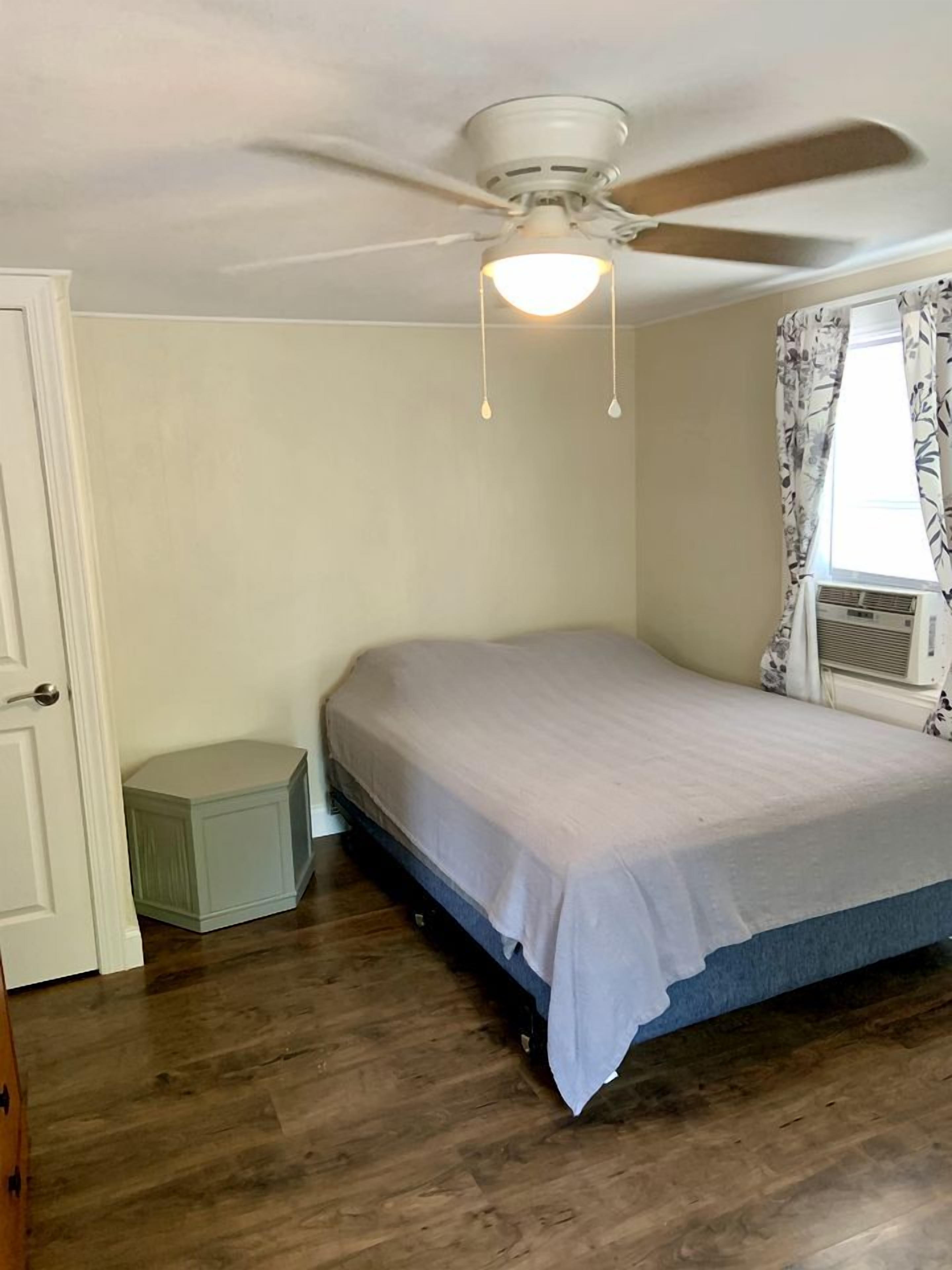Un dormitorio con una cama, ventilador de techo, ventana con cortinas y un pequeño armario blanco.
