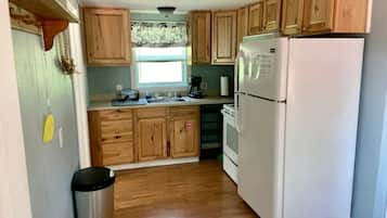 Una cocina con armarios de madera, un refrigerador blanco, un bote de basura de acero inoxidable y una ventana con cortina estampada.