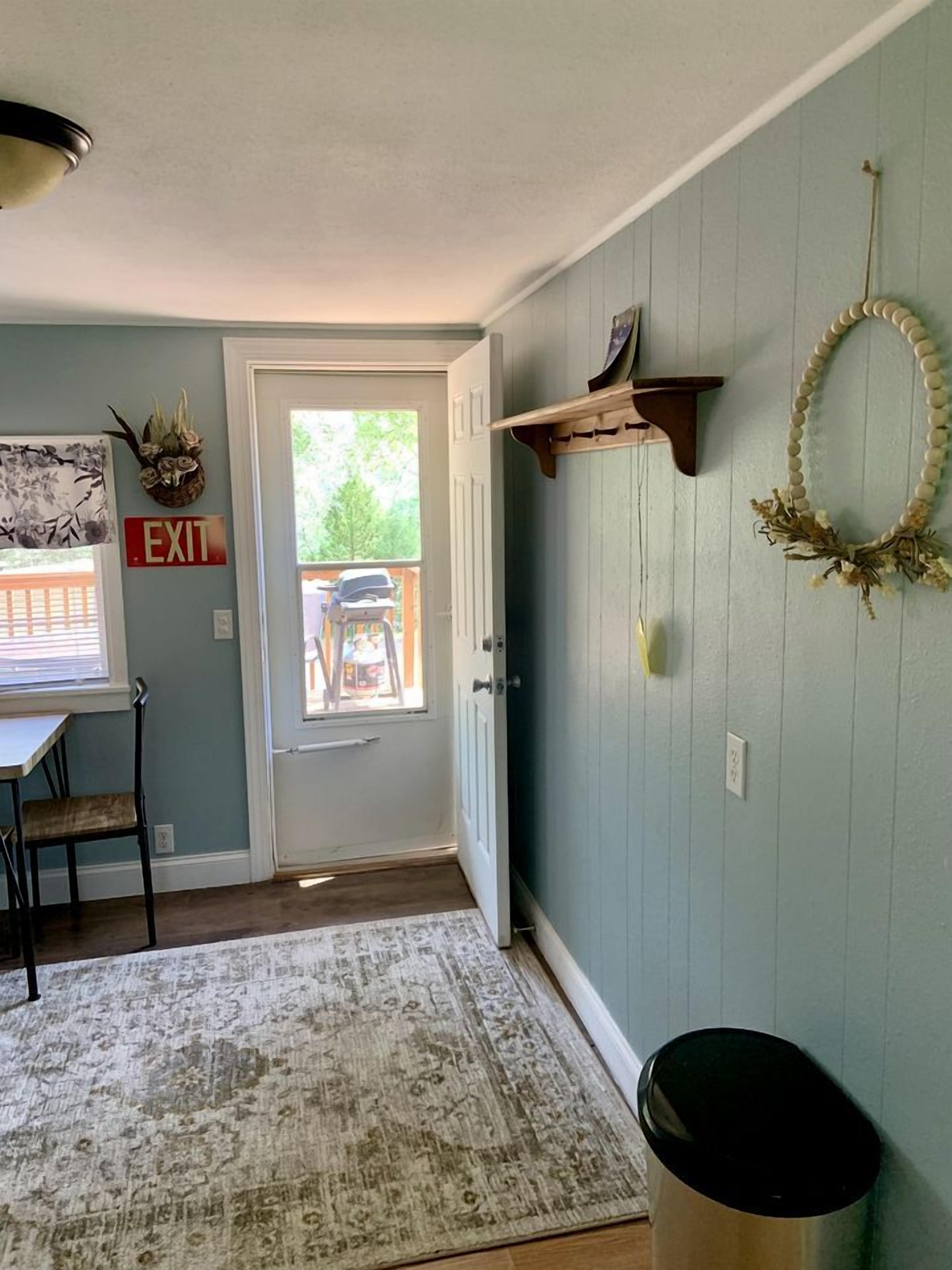 Una habitación con una puerta, una mesita con una silla, una alfombra, un cubo de basura y una pared con un adorno y una señal de salida.