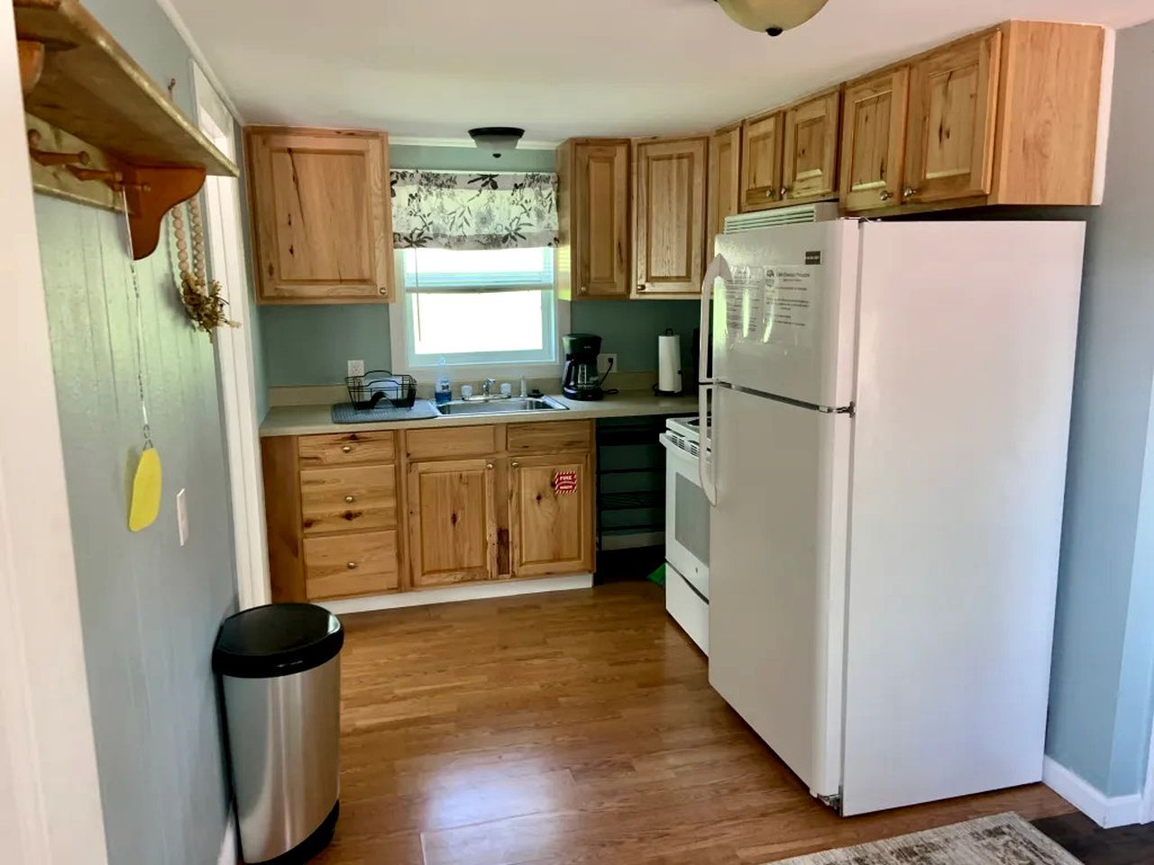 Una cocina con armarios de madera, un refrigerador blanco, un bote de basura de acero inoxidable y una ventana con cortina estampada.