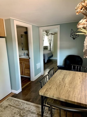 A dining area with a wooden table and chairs, a kitchen with cabinets and a sink, a bedroom with a bed and window, and a ceiling fan.