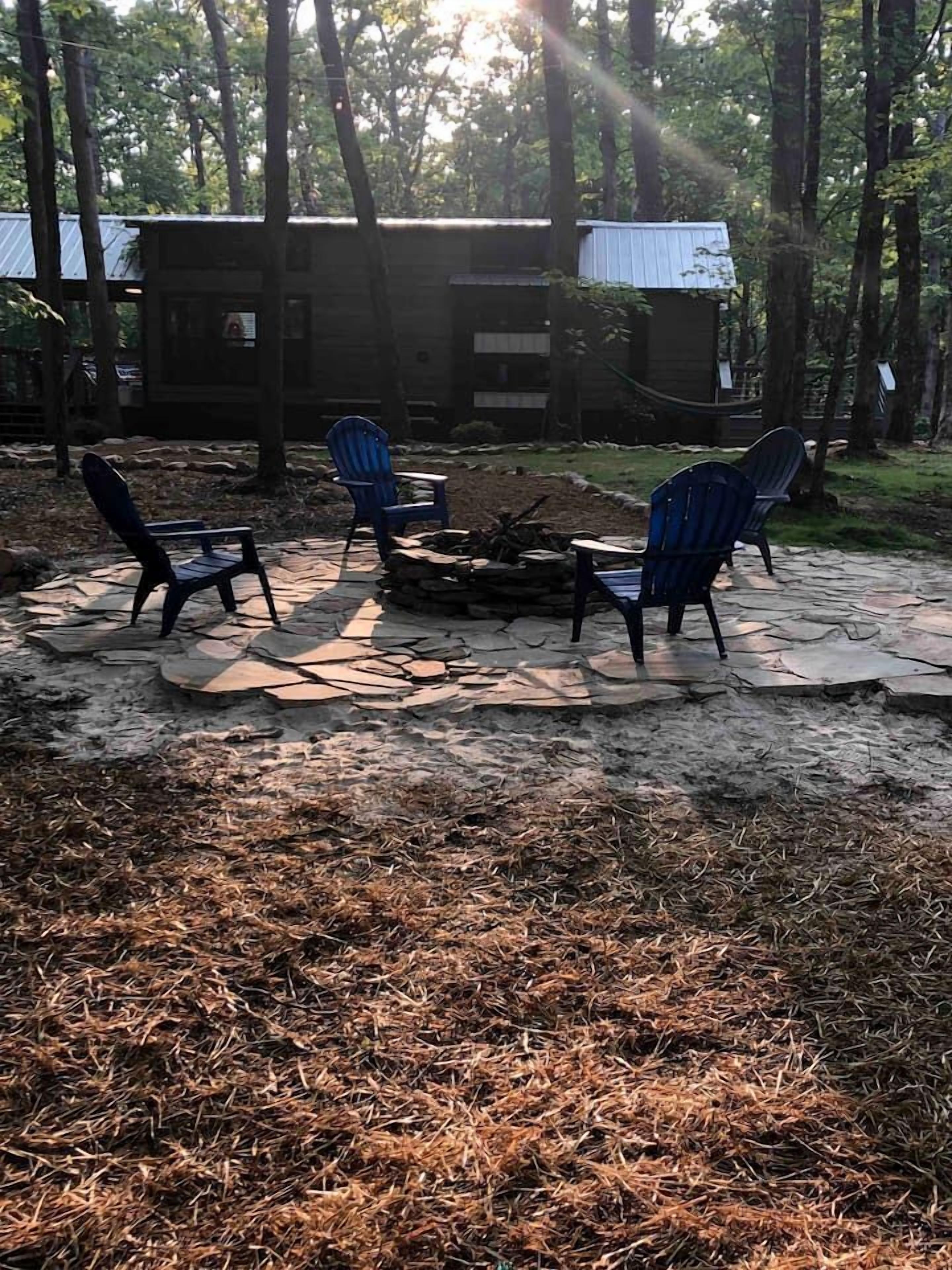 Una fogata rodeada de sillas Adirondack en un área boscosa con una cabaña al fondo.