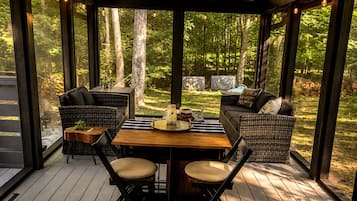 A covered outdoor seating area with wicker furniture, a wooden table, and a view of the forest.