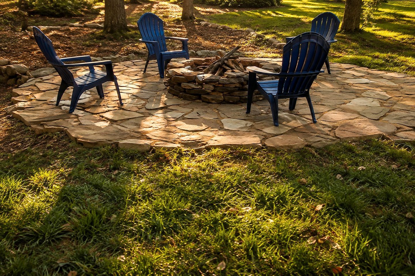 A stone fire pit surrounded by four Adirondack chairs in a garden setting.