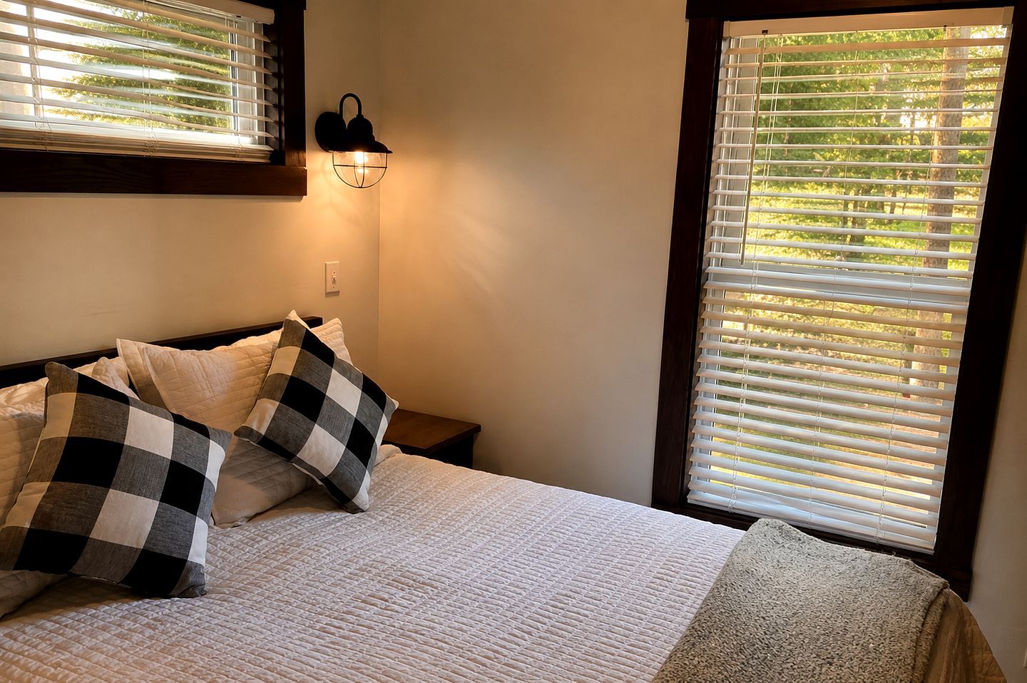 Un dormitorio con una cama, almohadillas de cuadros, una lámpara de pared y un ventanal con persianas.