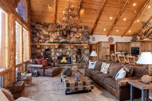 A cozy living room with a stone fireplace, wooden ceiling beams, and a chandelier. - Outstanding Lake Cabin with Hot-Tub, Views and Game Room in California (Big Bear Lake)