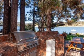 An outdoor grill area with stone walls, a wooden table, and a view of a lake. - Outstanding Lake Cabin with Hot-Tub, Views and Game Room in California (Big Bear Lake)