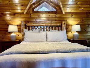 Une chambre à coucher dans une cabane en bois, avec un plafond en pente, un lit recouvert d’une couverture à motifs, deux lampes de chevet et une fenêtre.