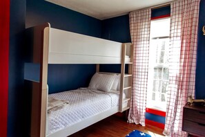 Ein Stockbett in einem Zimmer mit blauen Wänden, einem Fenster mit Karottenvorhängen und einer kleinen Holzkommode.