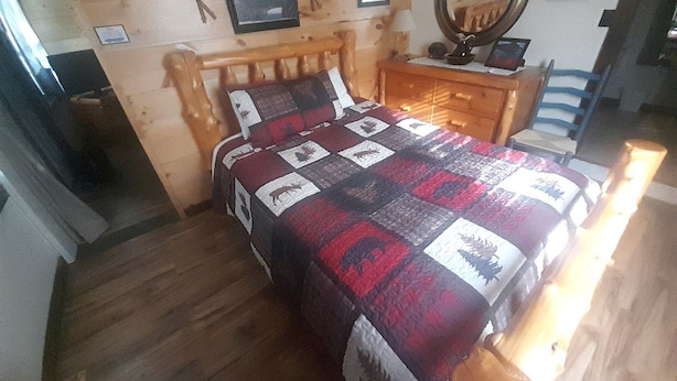 Puinen sänky, jossa on peitto, puinen lipasto, peili ja seinällä oleva kehystetty kuva.