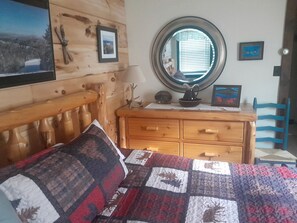 Ein Schlafzimmer mit einem Holzkopfteil, einem Bett mit Tagesdecke, einer Kommode mit Schubladen, einem runden Spiegel und einem Stuhl.