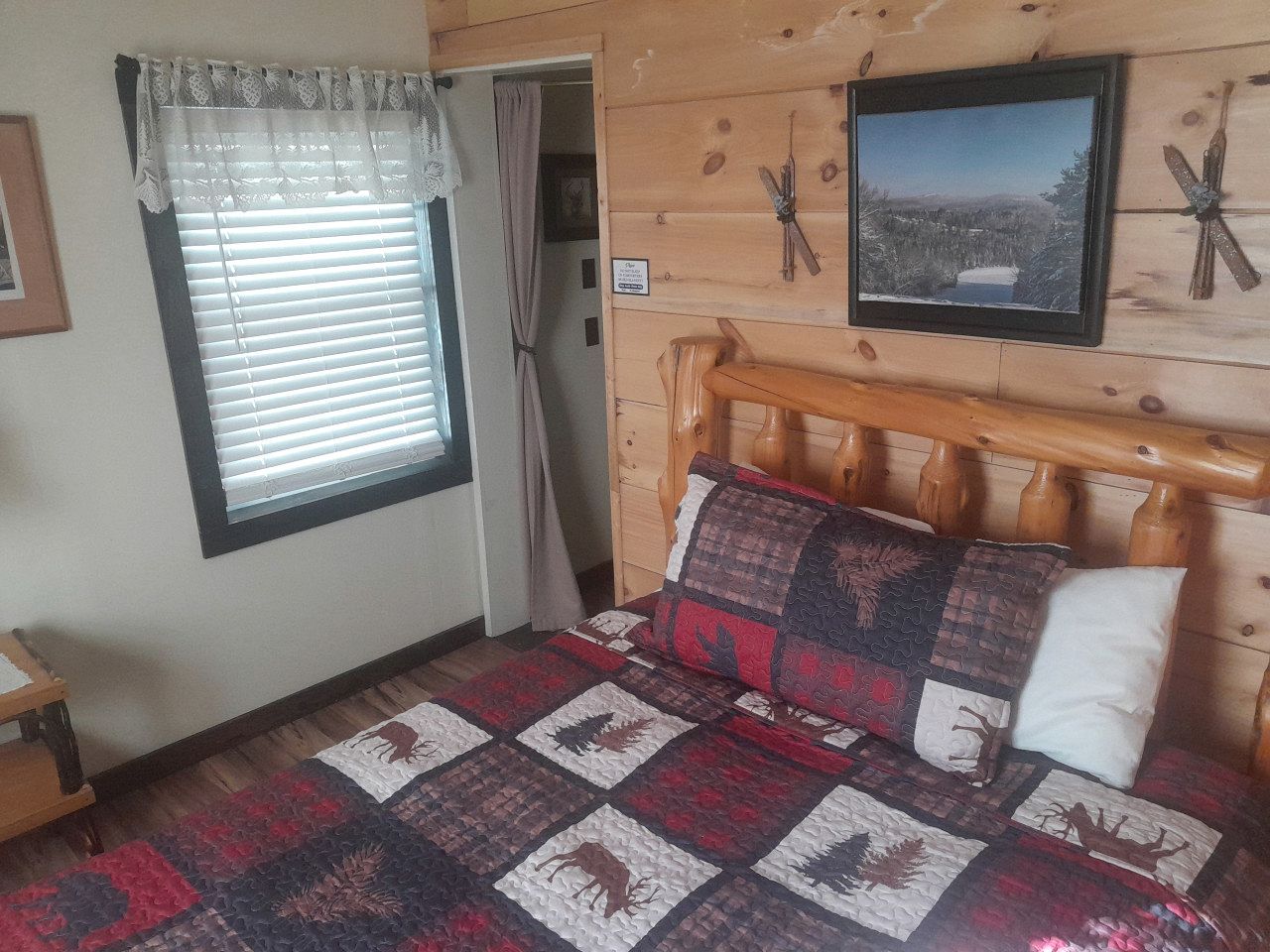 A wooden cabin room with a bed, a window with blinds, a door, and a wall-mounted picture.