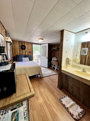 Een kamer met een bed, een bureau met een computer, een houten vloer, een badkamer met een wastafel en spiegel, en een deur die naar buiten leidt.