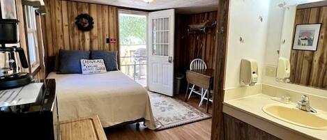 A room with a bed, a desk with a computer, a wooden floor, a bathroom with a sink and mirror, and a door leading outside.