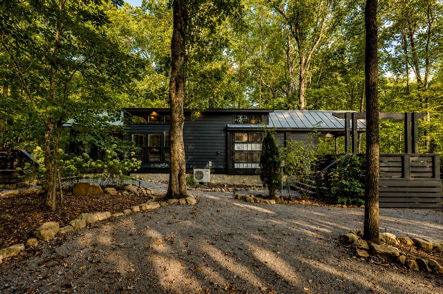 A modern cabin with a metal roof, surrounded by a natural landscape with trees and a gravel driveway.