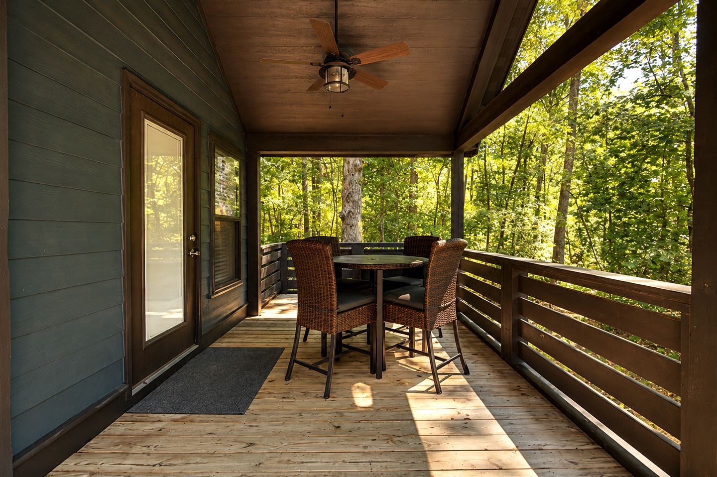 A covered outdoor area with a table and chairs, a ceiling fan, and a view of a forest.