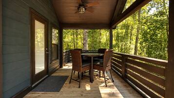 A covered outdoor area with a table and chairs, a ceiling fan, and a view of a forest.