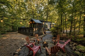 Un rustico chalet con una veranda, immerso in una foresta. Si notano poltrone in stile Adirondack e un braciere.